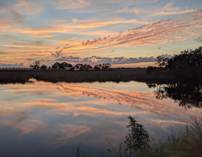 Photo picturing a sunset reflected in a large inlet, surrounded by marsh