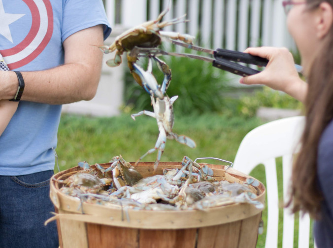 Woman outdoors removing blue crabs from a bushel basket with tongs